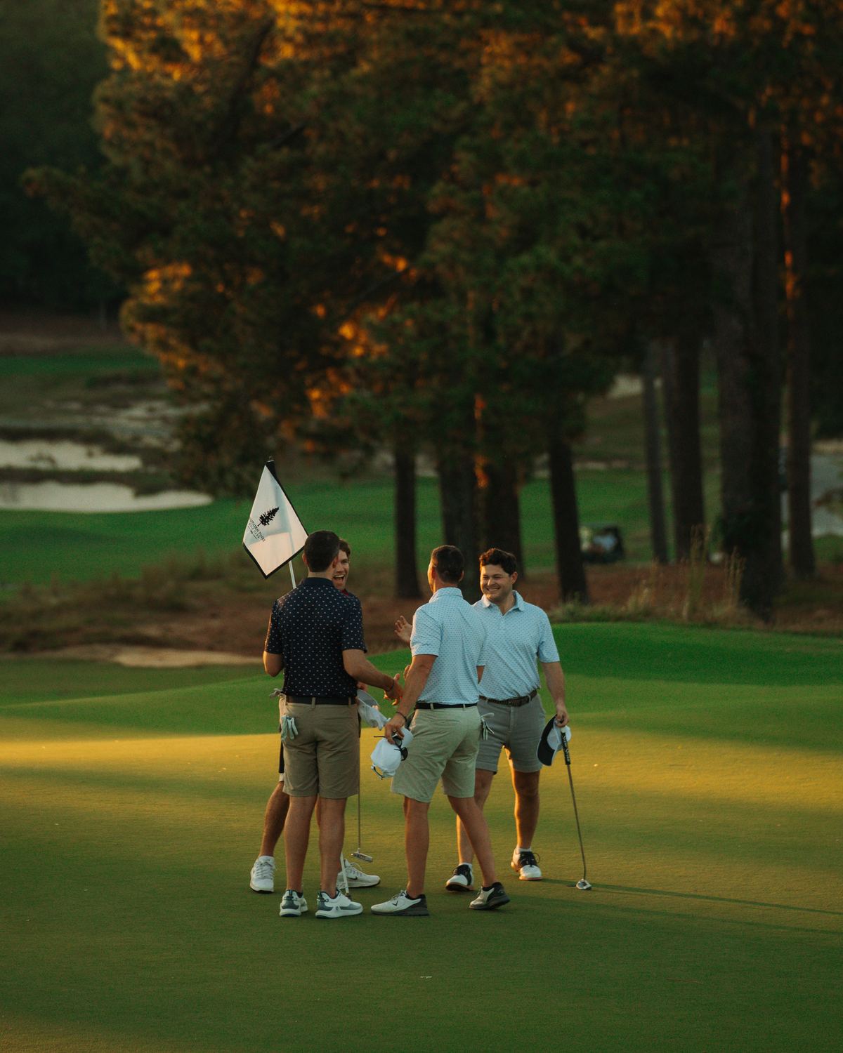 Four golfers shake hands beside a white pin flag on the green at sunset, tall pines glowing gold behind them