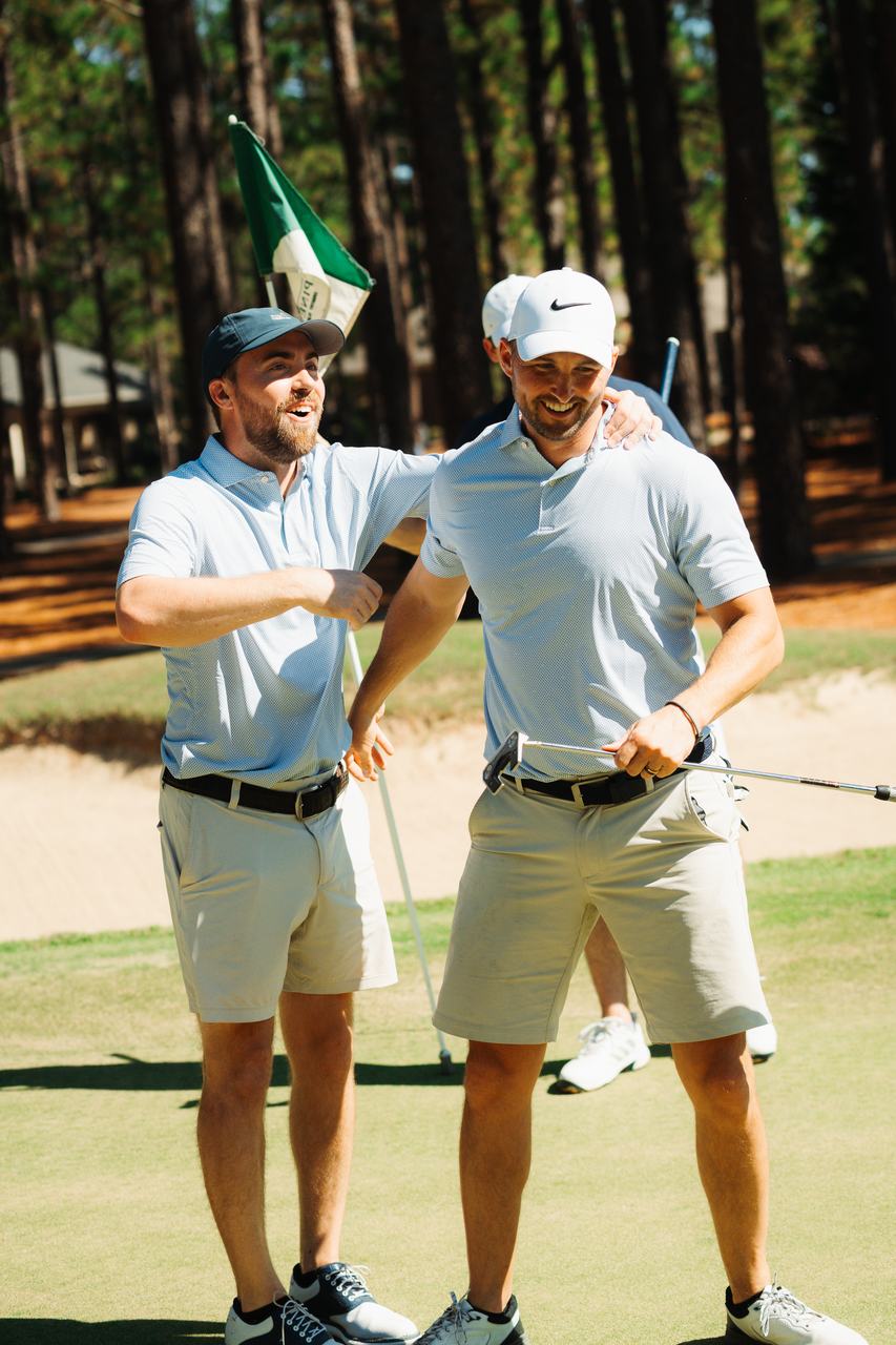 Two smiling golfers in light blue polos embrace on the green beside a green pin flag, a third partly visible behind, warm sunlight