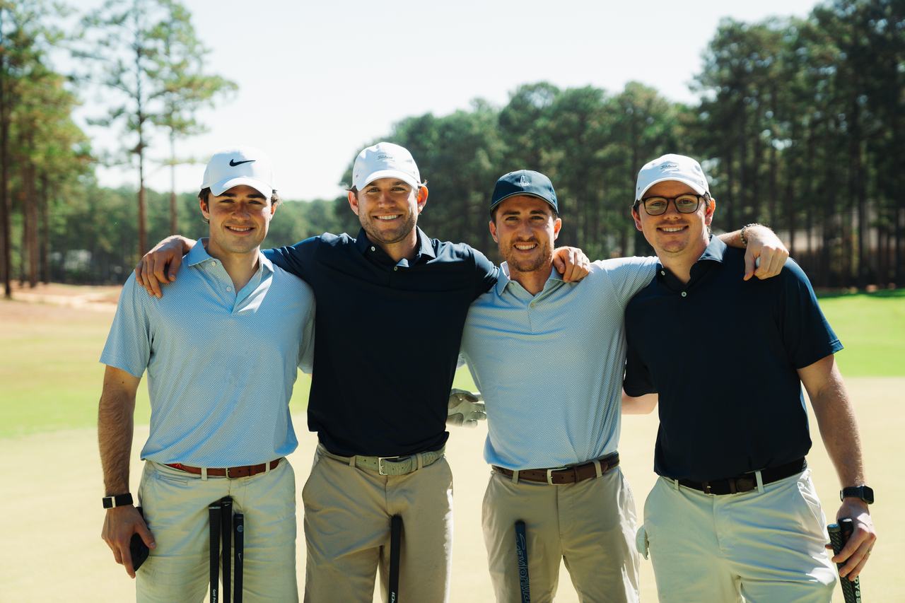 Four smiling golfers in light blue and navy polos stand arm in arm on a sunny fairway at Pinewild Country Club