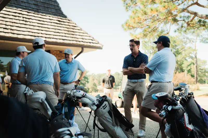 A group in light blue polos and caps gathers beside golf bags outside a shingle-roofed clubhouse, two men with arms crossed talking at right, warm afternoon light.