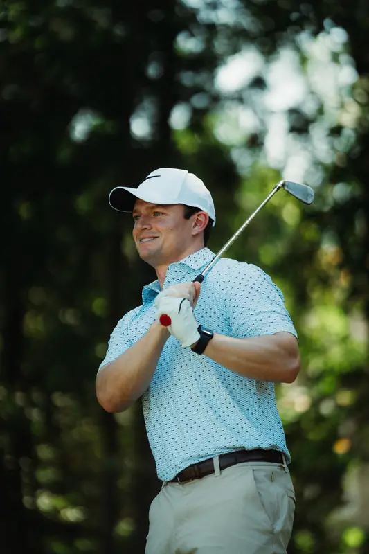 A smiling golfer in a white cap, light blue polo and black watch holds his iron across his body, enjoying the moment, dappled green background.