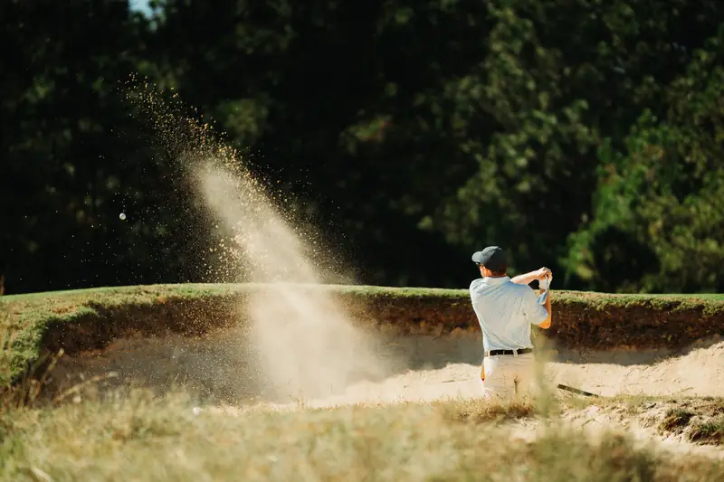 Golfer in light blue polo and dark cap blasts out of a sand bunker, a plume of golden sand backlit against dark trees.