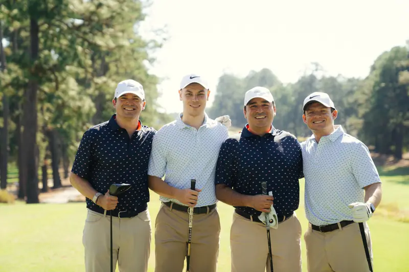 Landscape group portrait of four grinning golfers in white caps, navy and light polos with clubs, arms around shoulders in golden sun.