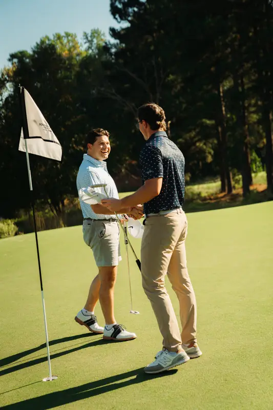Two golfers, one in a light blue polo laughing and one in a navy polo seen from behind, shake hands on the green beside a flag in golden light.