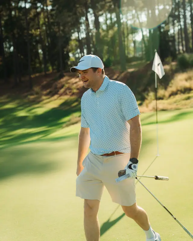 A golfer in a light blue patterned polo and white cap laughs mid-stride on a sunlit green, club and glove in hand, flag and warm pines behind.