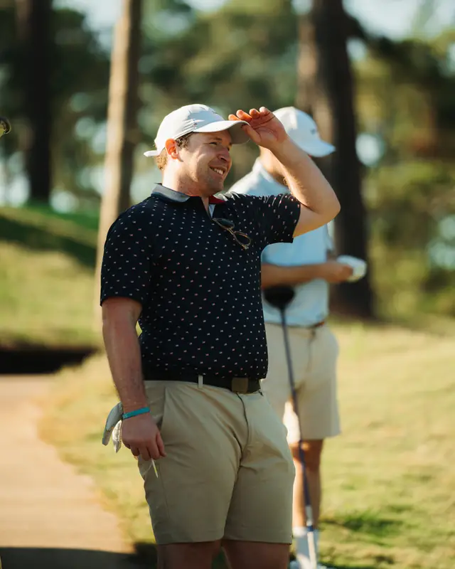 Smiling man in a white cap and navy patterned polo shields his eyes from the sun, sunglasses hooked on his collar, another golfer blurred behind.