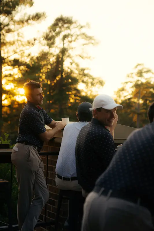 Golden-hour terrace moment: a man in a navy dotted polo leans on the rail with a cup while a smiling man in a white cap glances back, sun flaring behind the trees.