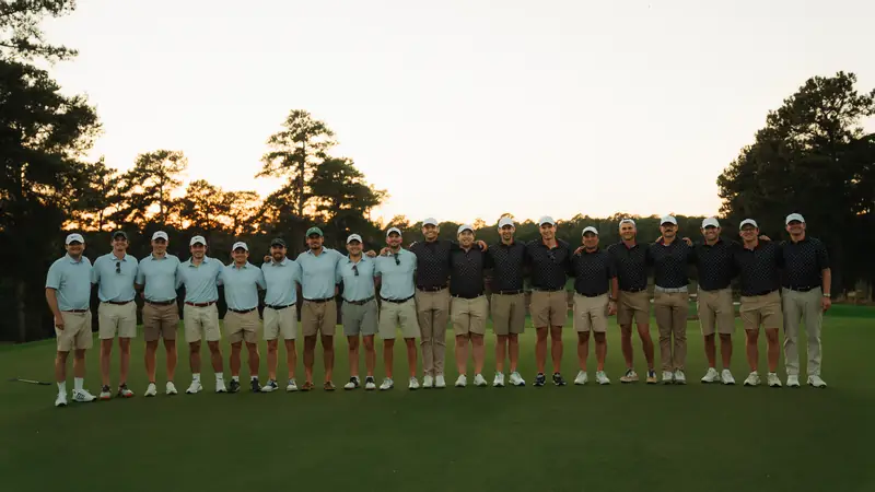 Two teams in light blue and navy polos stand arm in arm in one long row on the green at golden-hour dusk.