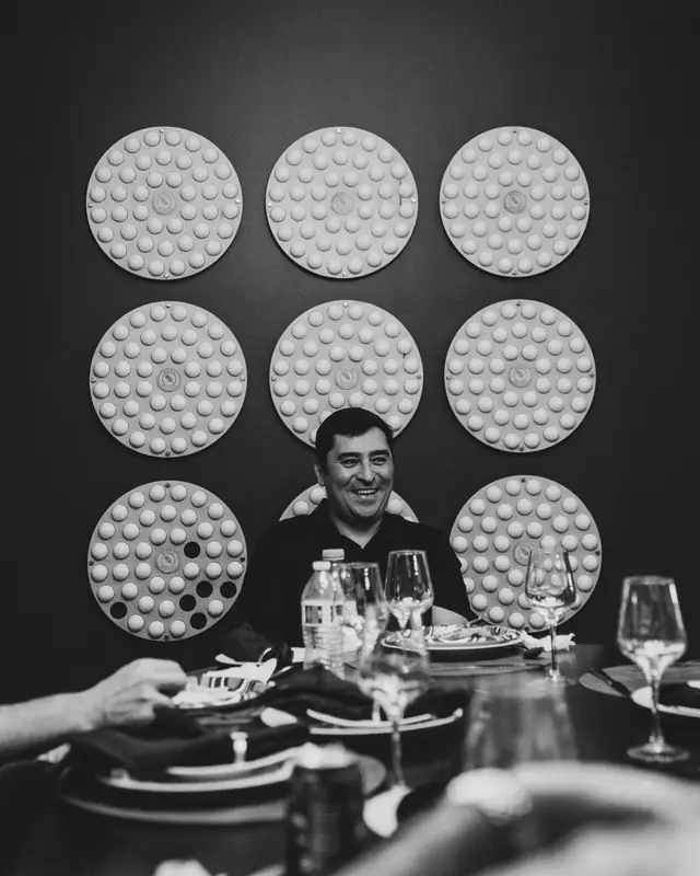 Black-and-white portrait of a man laughing broadly at a dinner table, framed by rows of round wall plates and wine glasses.
