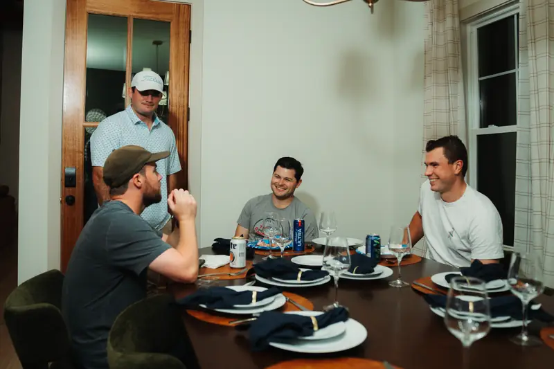 Around a dinner table set with navy napkins, three men laugh warmly while a fourth in a cap and green hat stands behind.