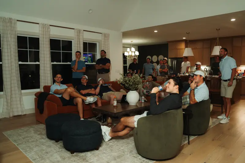 A big group of men in golf polos and caps gathers in a warm living room and kitchen, smiling and watching something off-camera, one drinking from a can.
