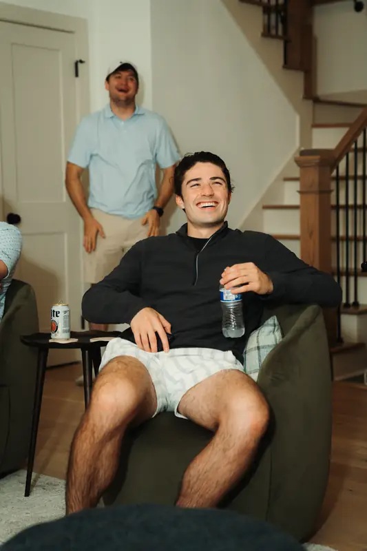 A young man in a black quarter-zip and plaid shorts laughs openly in an olive armchair holding a water bottle, with a grinning man in a cap behind him.