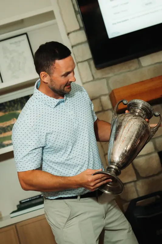 A man in a light blue patterned polo holds up an antique silver trophy cup beside a stone fireplace, looking at it with a slight smile in warm indoor light.