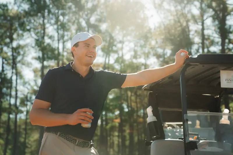 Laughing golfer in navy polo and white cap holds a drink can and leans on a golf cart, backlit by golden sun through pines.