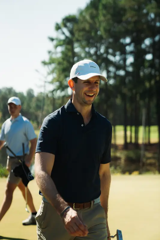 Close portrait of a laughing golfer in white cap and navy polo walking on the course, a companion behind him.