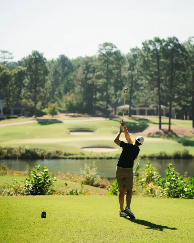 Golfer in navy polo and white cap finishes a tee shot over a pond toward a fairway with bunkers in warm light.