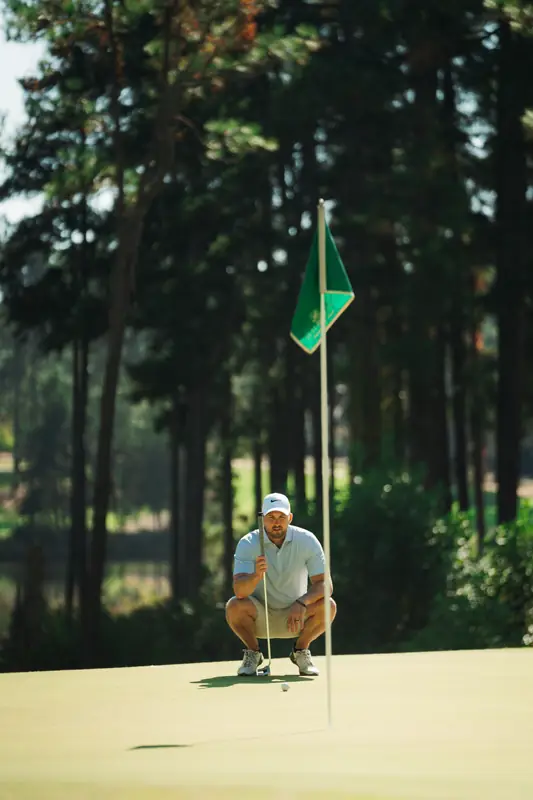 Golfer in light polo and white cap crouched on the green reading a putt beside a green flag, tall pines behind in warm light.