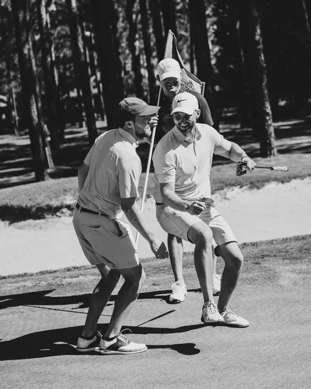 Black and white frame of three golfers laughing and dancing playfully around the pin on the green, one holding a putter.