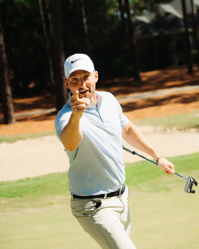 A golfer in a white cap and light polo points at the camera with an excited grin, putter in hand, sand bunker behind.
