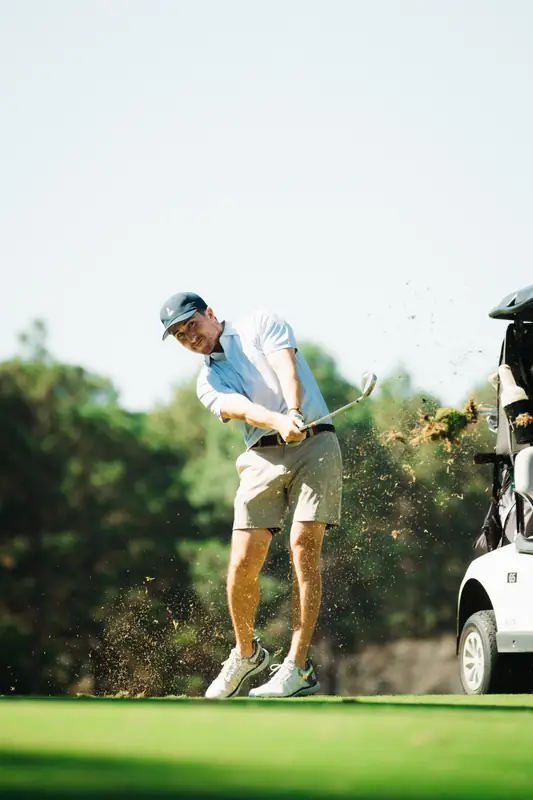 Golfer in light blue polo and navy cap smiles through the swing as turf and dirt fly in golden light, golf cart at right.