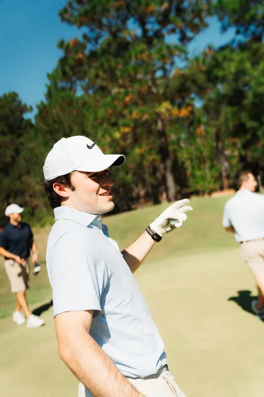 Close portrait of a smiling golfer in white cap and light blue polo pointing down the course with a glove, two other players softly blurred behind.