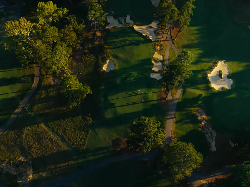 Top-down drone shot of a fairway and green with four tiny golfers, long shadows, bunkers and a cart path.